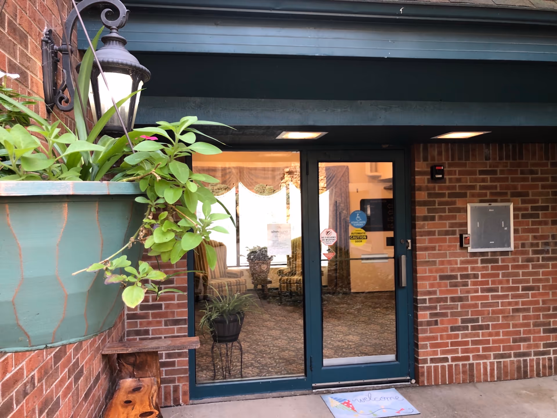 Glass entrance doors to a brick senior living facility with a hanging planter and a welcome mat.