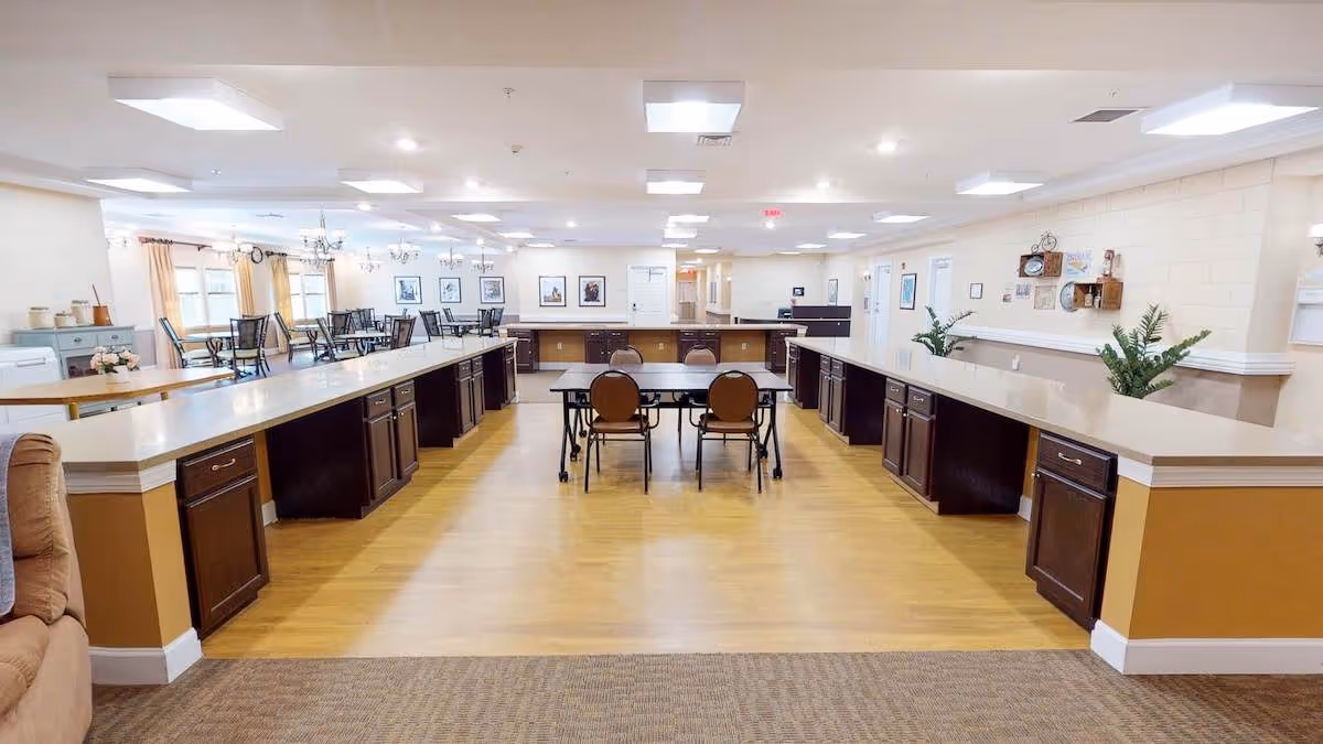 A spacious, well-lit common area with long countertops and dark wood cabinets on both sides, a small table with four chairs in the center, and a dining area with tables and chairs in the background. The room has light-colored walls, wooden flooring, and ceiling lights.