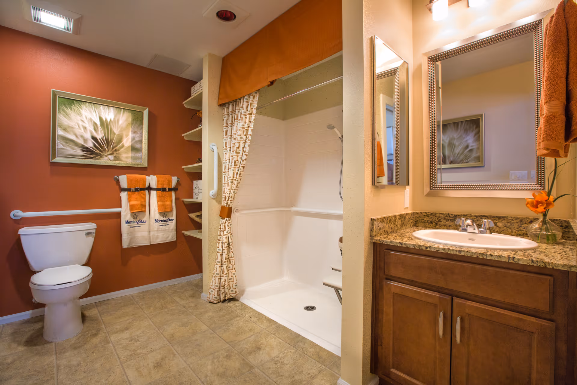A bathroom with a toilet, a walk-in shower with a patterned curtain, a sink with a granite countertop and wooden cabinet, a large mirror, orange towels hanging on a rack, and a framed floral artwork on the wall.