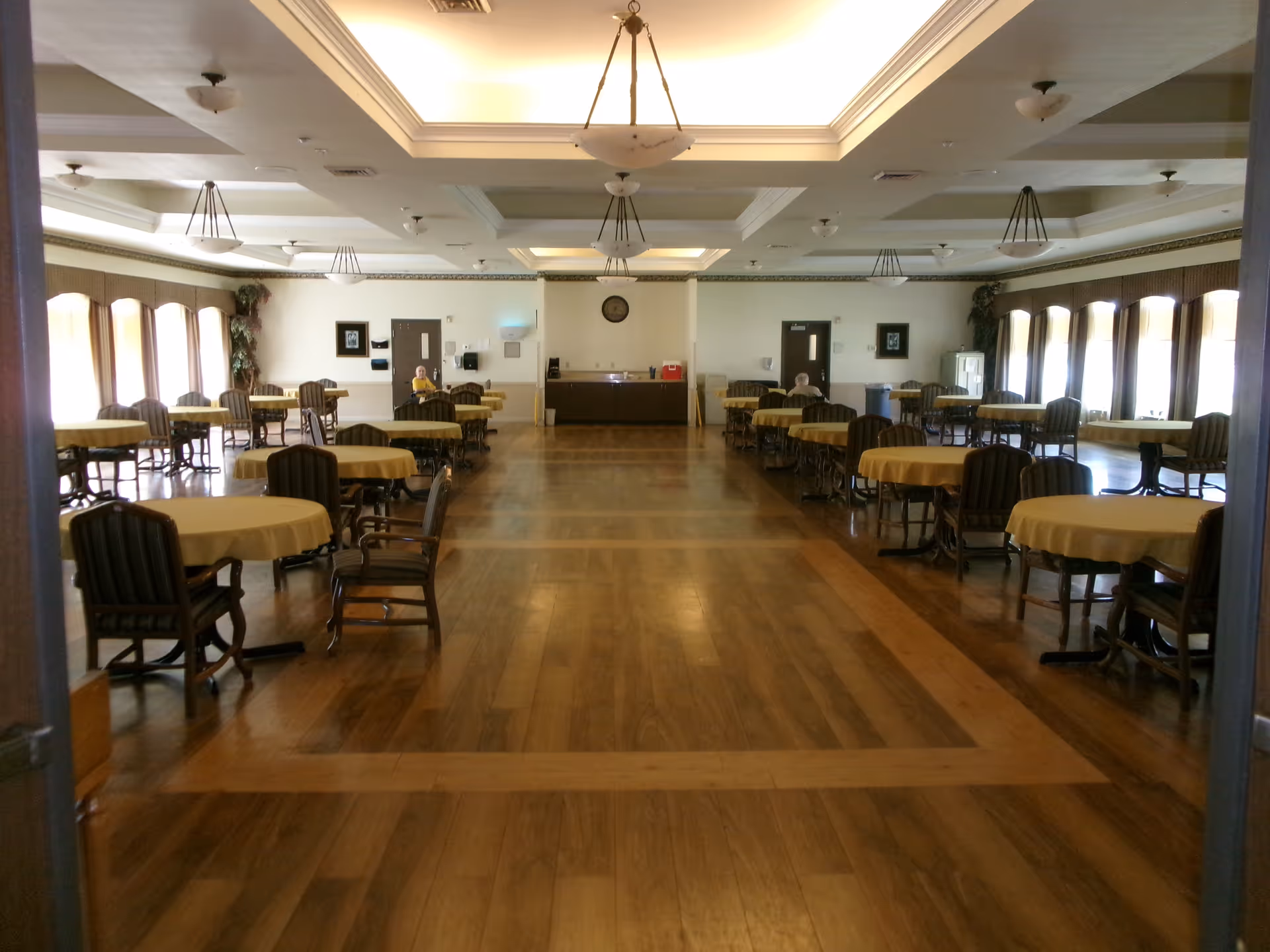 Large dining room with round tables covered in yellow tablecloths and wooden chairs arranged on a polished wood floor.