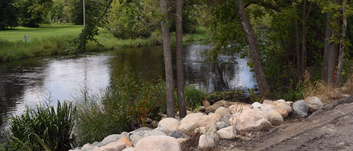 A peaceful outdoor scene featuring a calm river bordered by trees and bushes, with a rocky embankment and a dirt path alongside the water.