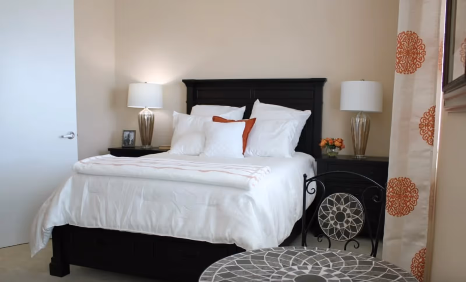 Bright bedroom featuring a dark wood bed with white bedding, matching bedside tables and lamps, patterned curtains, and a small round mosaic table in the foreground.