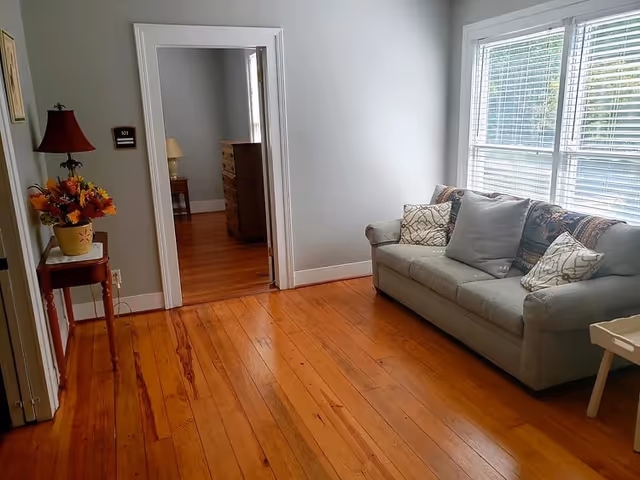 Bright living room with hardwood floors, a sofa by a large window, and a doorway leading to another room with a dresser.
