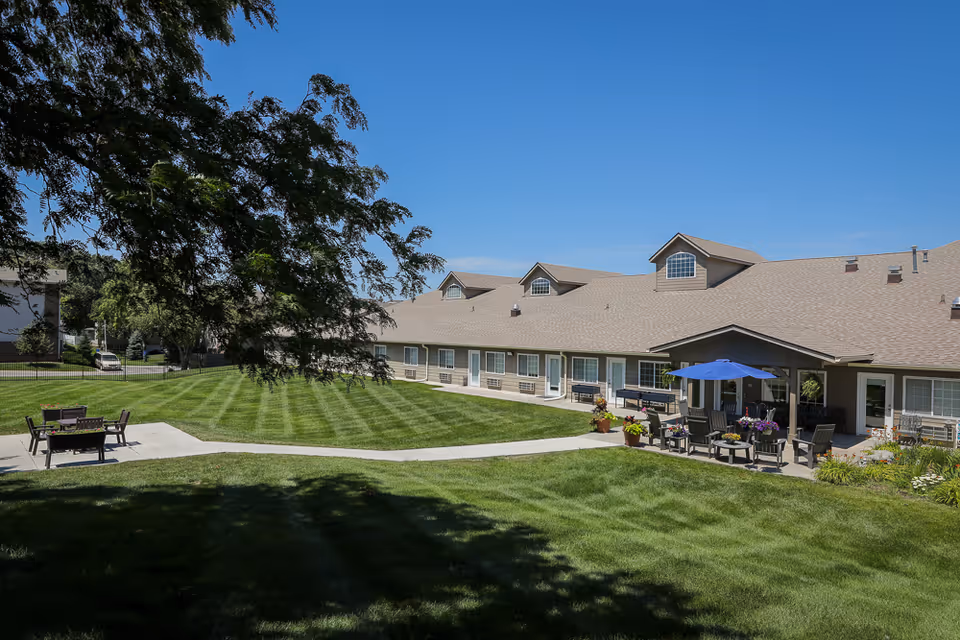 Single-story assisted living building with a manicured lawn, patio seating and a blue umbrella in a sunny courtyard.