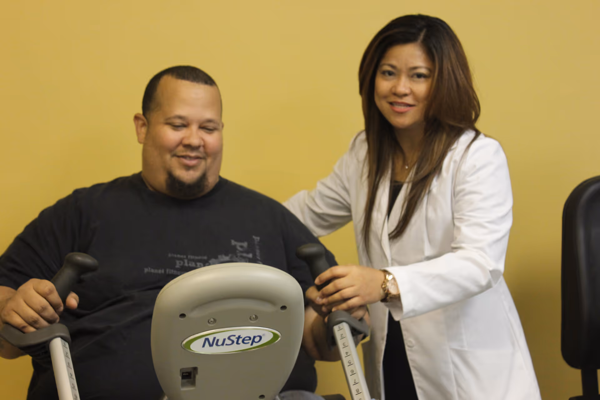 A man is using a NuStep exercise machine while a woman in a white coat, possibly a healthcare professional, stands beside him smiling and providing support.