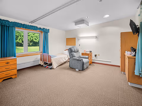 A spacious senior living bedroom with a single bed next to a window with blue curtains. The room includes a gray recliner chair, wooden dressers, a wall-mounted TV, and beige carpet flooring. The walls are white with minimal decorations.