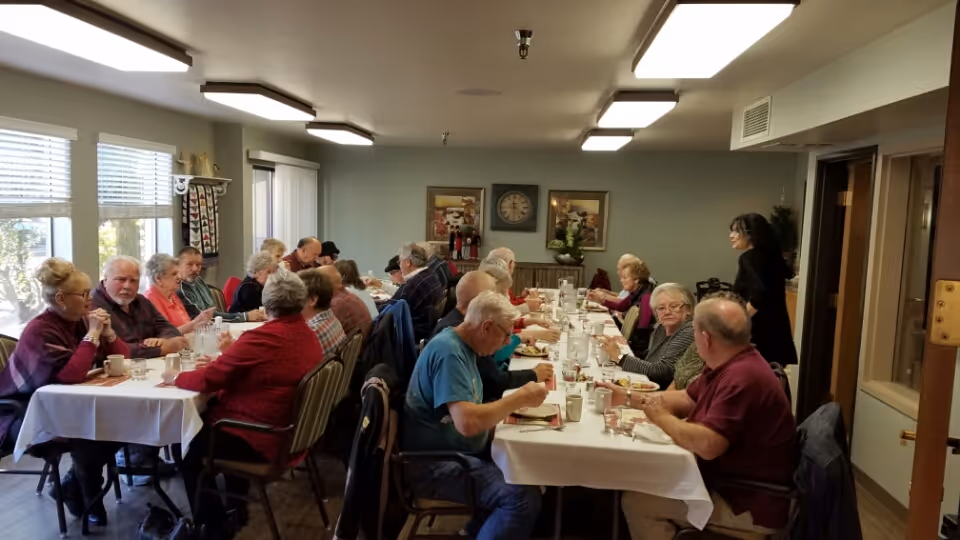 A group of elderly people sitting around two long tables covered with white tablecloths, eating and socializing in a well-lit dining room with large windows and framed artwork on the walls.