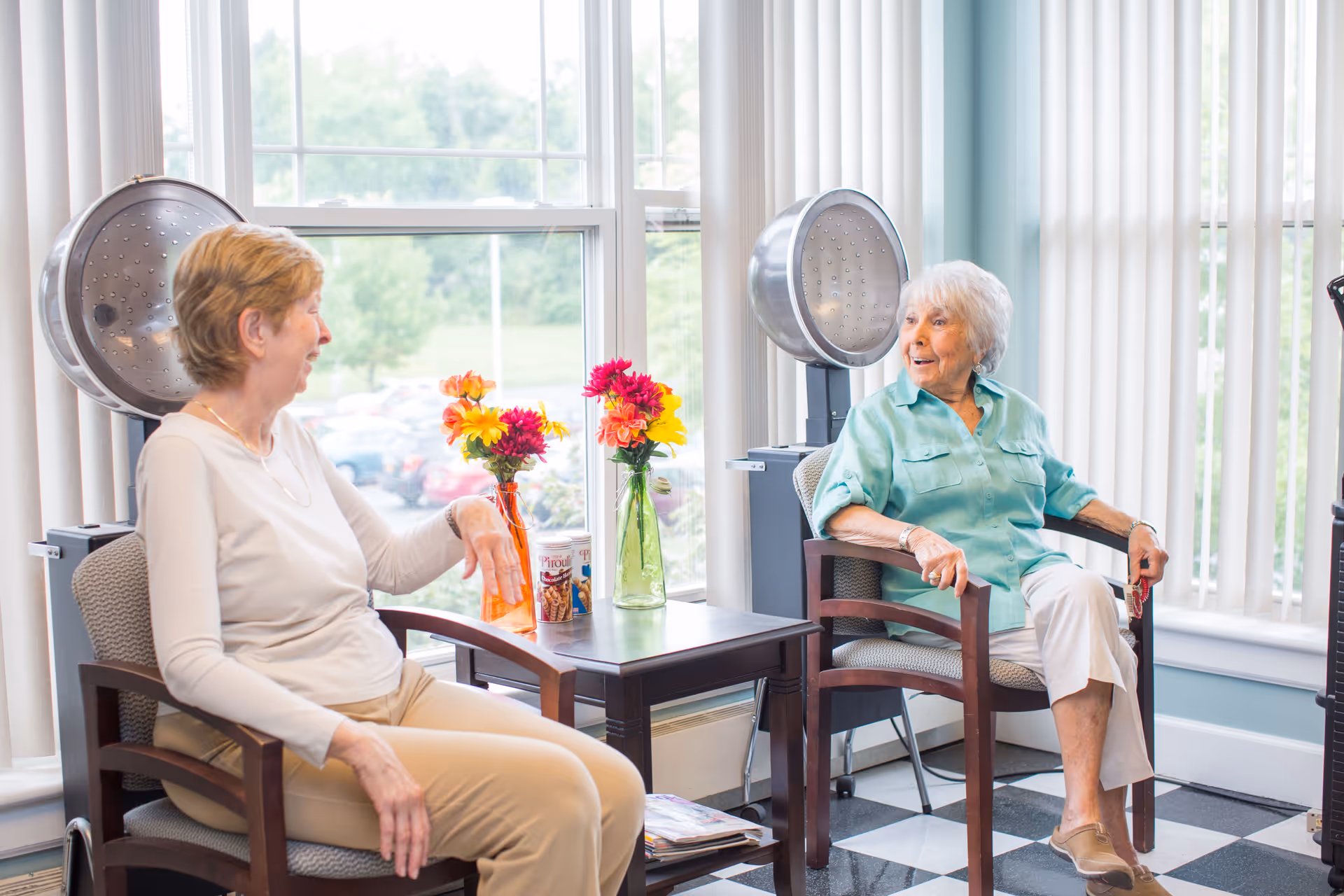 Two elderly women sitting and chatting in a bright room with large windows and vertical blinds. They are seated in wooden chairs with cushions, and there are two vintage hair dryers behind them. A small table between them holds two vases with colorful flowers and a container of snacks. The floor has a black and white checkered pattern.