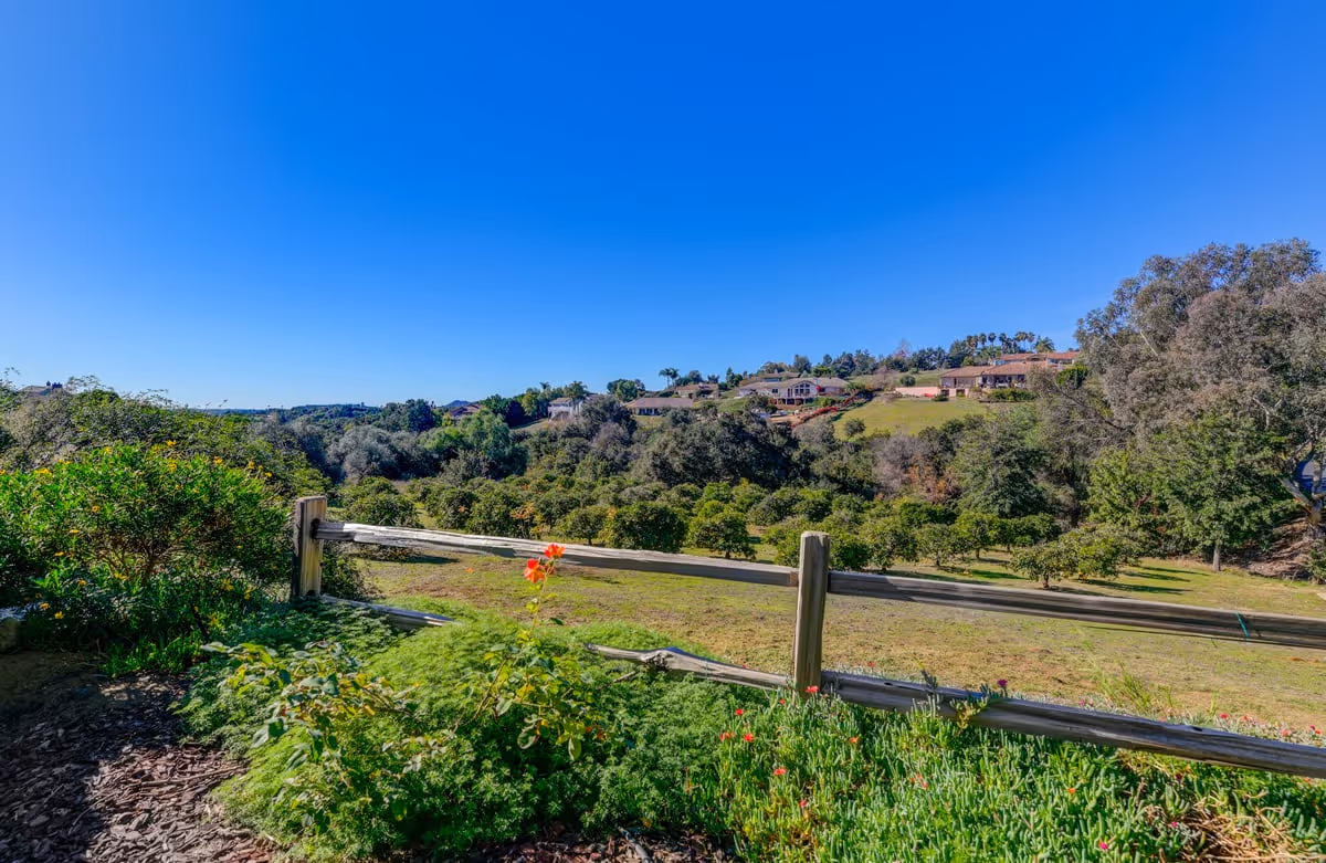 A scenic outdoor view of a garden area with green shrubs and flowers in the foreground, a wooden fence, and a landscape of trees and houses on rolling hills under a clear blue sky.