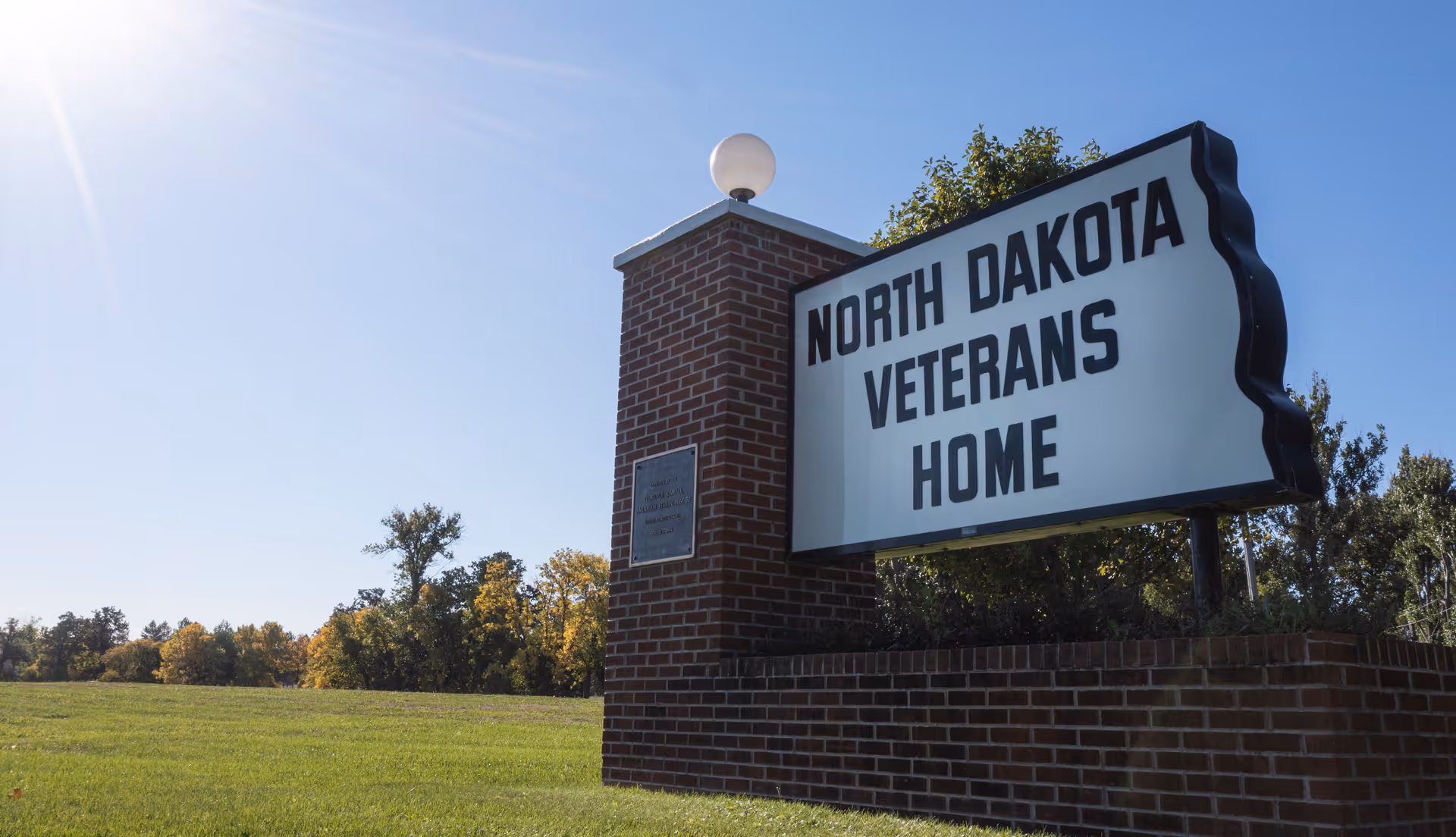 A large brick signpost with a white sign reading 'NORTH DAKOTA VETERANS HOME' situated on a grassy area with trees and a clear blue sky in the background.
