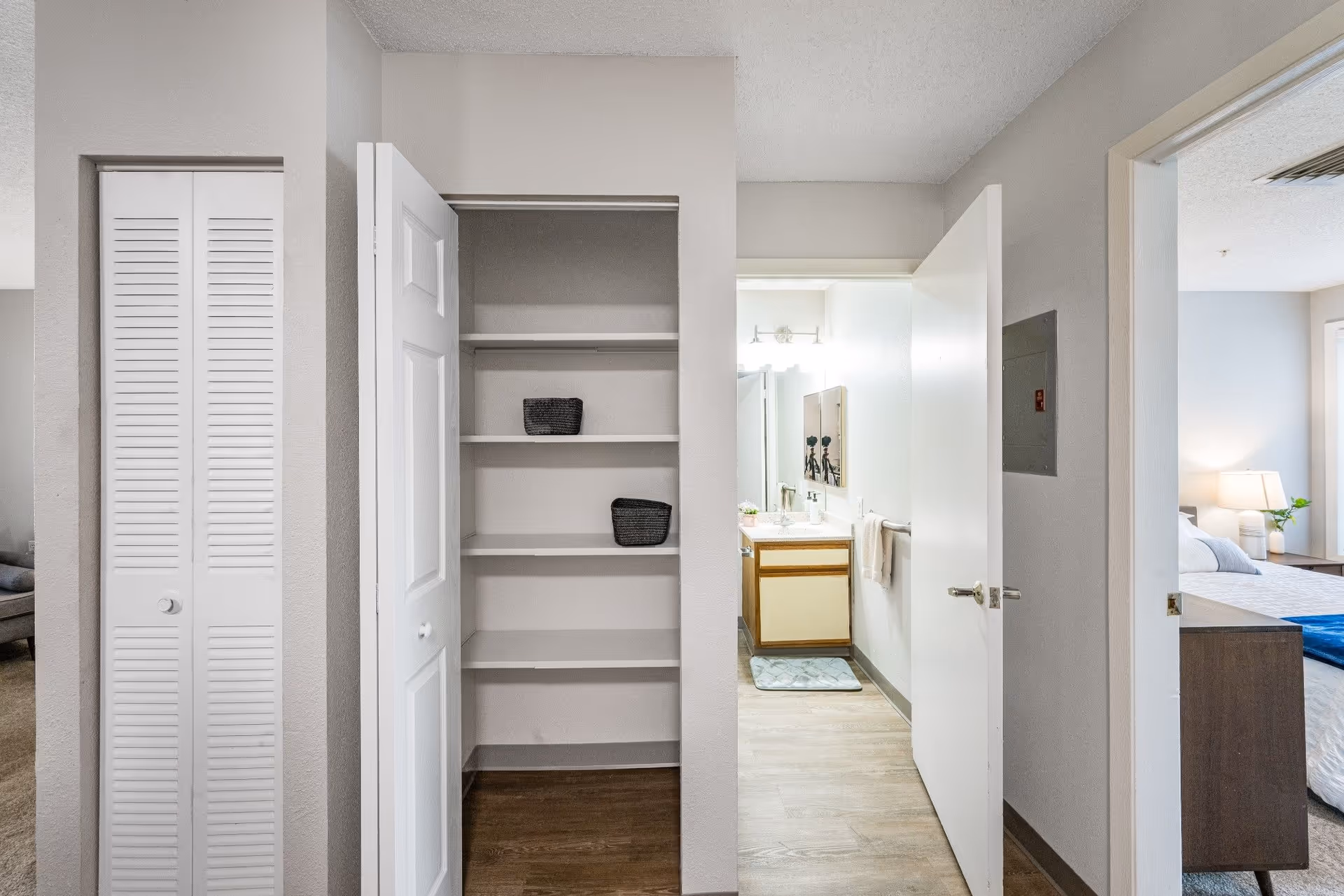 View of an interior hallway area showing an open closet with shelves and two small baskets, a partially open door leading to a bathroom with a sink and mirror, and a glimpse of a bedroom with a bed, nightstand, lamp, and window.
