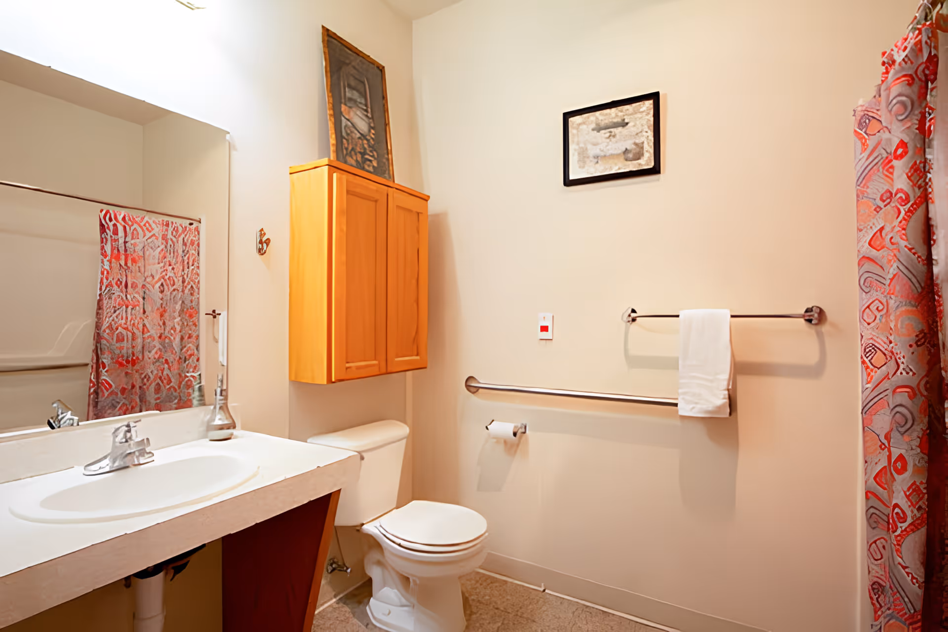 A bathroom with a white sink and countertop, a toilet, a wooden cabinet mounted on the wall above the toilet, a towel rack with a white towel, a grab bar, a toilet paper holder, and a shower with a red and white patterned curtain. There are two framed pictures on the walls.