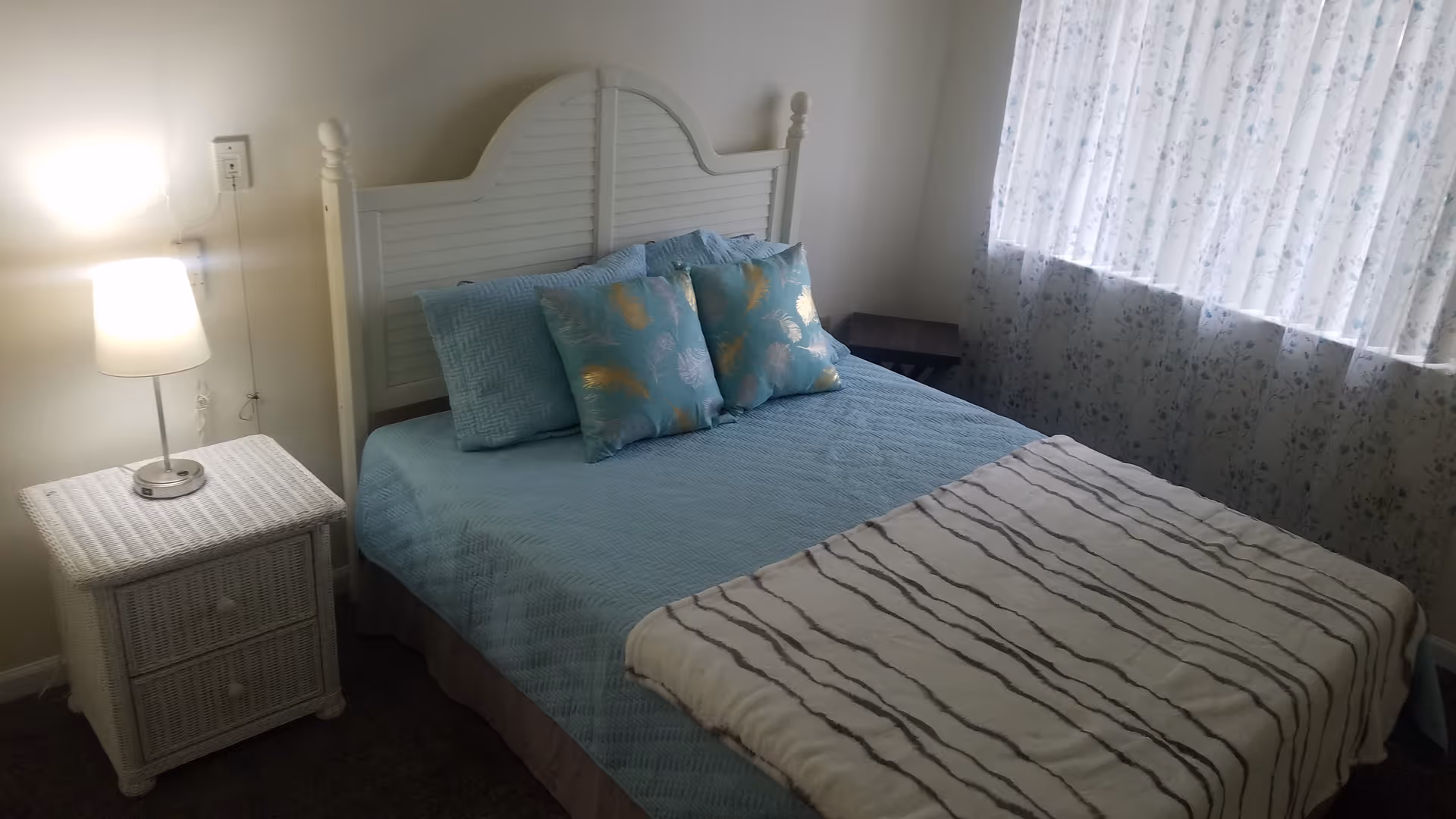 Cozy bedroom with a white wooden headboard, blue bedding and decorative pillows, a bedside table with a lamp, and a curtained window.