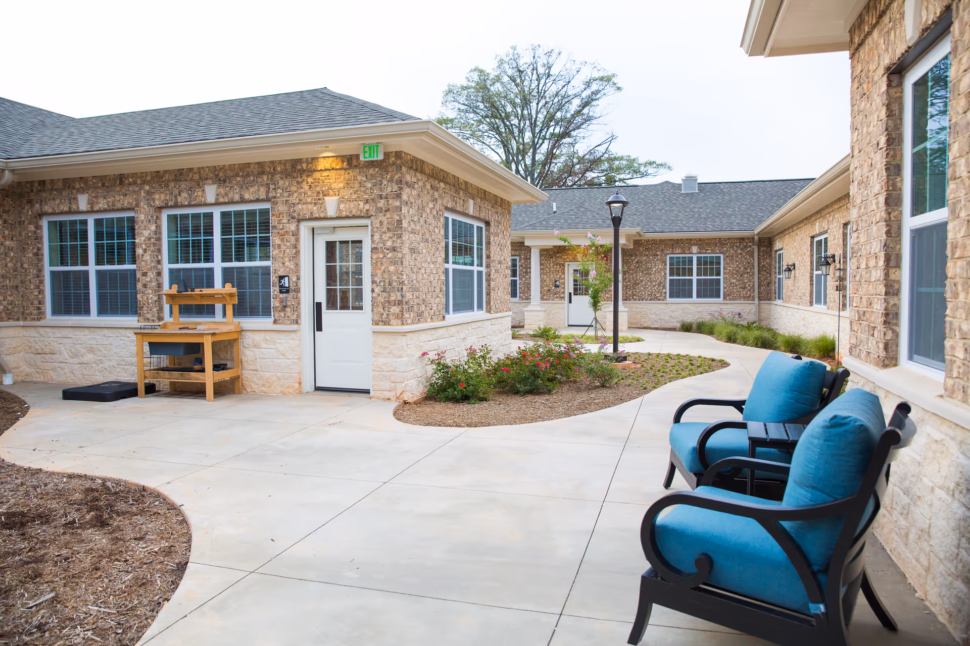 Brick courtyard of a senior living facility with a paved walkway, blue cushioned chairs, landscaping, and building entrances.