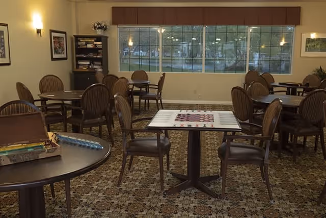 A well-lit common room with multiple round tables and wooden chairs arranged throughout the space. One table in the foreground has a checkerboard game set up. Large windows line the back wall, allowing natural light to enter. The room has patterned carpet flooring and framed artwork on the walls.