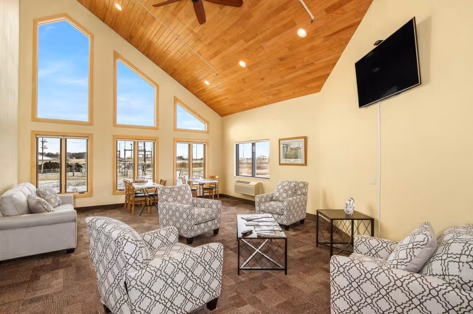 Spacious senior living common room with patterned armchairs and sofas arranged around coffee tables, a dining table near large windows, and a high wood-paneled ceiling.