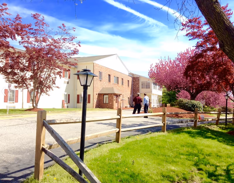 Exterior view of a senior living facility with a two-story building featuring white and brick walls. Two people are walking on the driveway near the building. The scene includes a wooden fence, green grass, and trees with red and pink blossoms under a blue sky with wispy clouds.