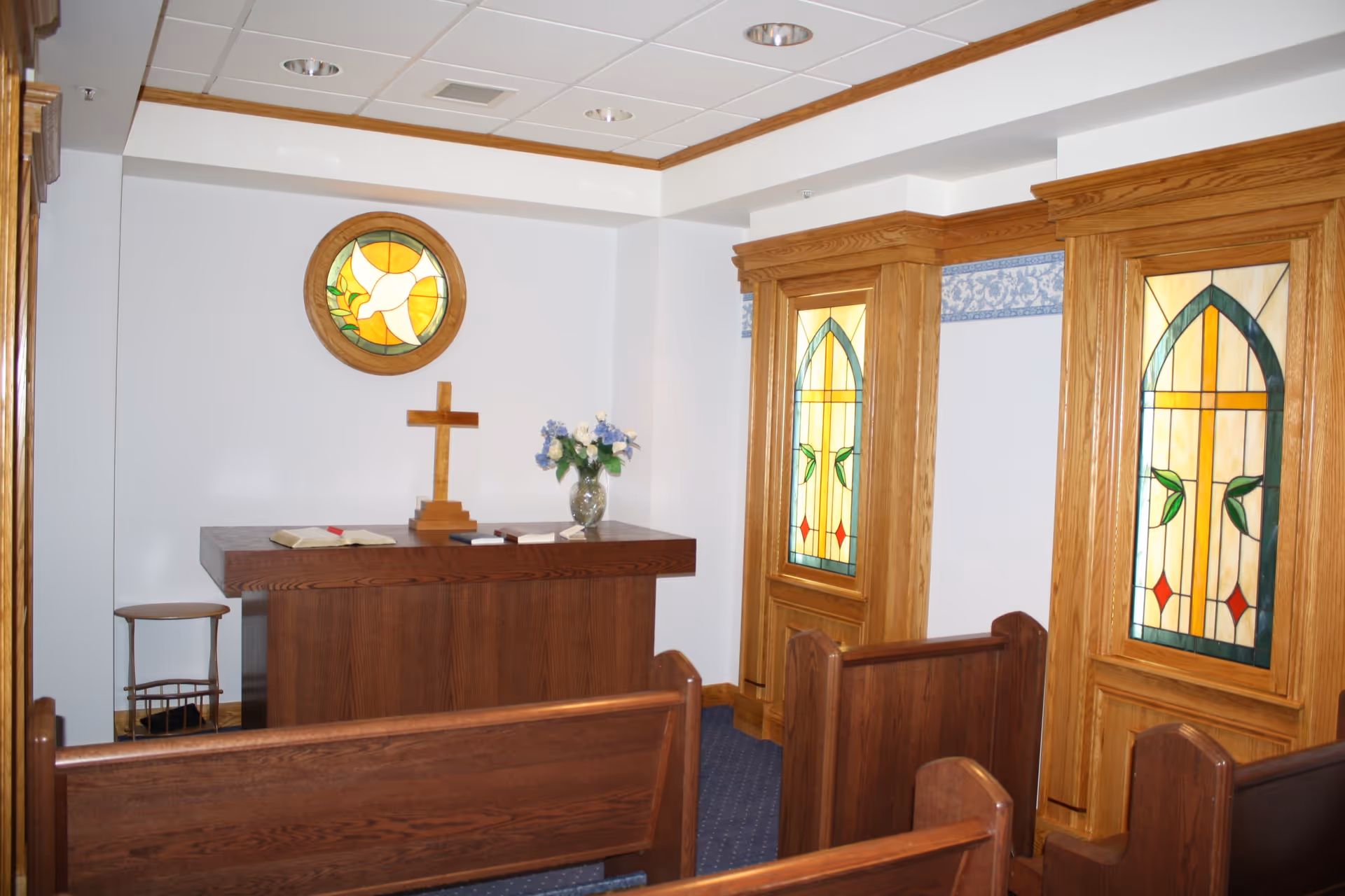 Small chapel room with wooden pews, a wooden altar with an open Bible, a wooden cross, and a vase of flowers. The room features stained glass windows with religious designs and a round stained glass window with a dove above the altar.