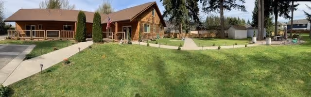A panoramic view of a senior living facility with a large green lawn in the foreground. The facility consists of a single-story wooden building with a brown roof and several windows. There are paved walkways leading to the building entrance, and tall trees and shrubs are visible around the property. The sky is clear with some clouds.