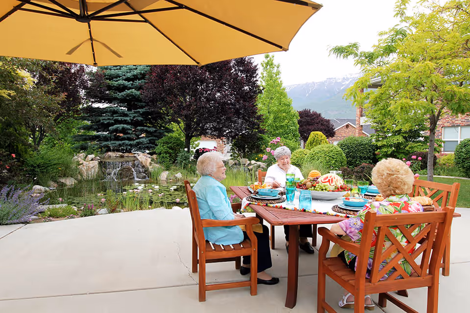 Three elderly women sitting around a wooden outdoor dining table under a large yellow umbrella, enjoying a meal with plates, glasses, and a fruit centerpiece. The setting is a well-maintained garden with lush greenery, flowering plants, a small pond with a waterfall, and mountains visible in the background.