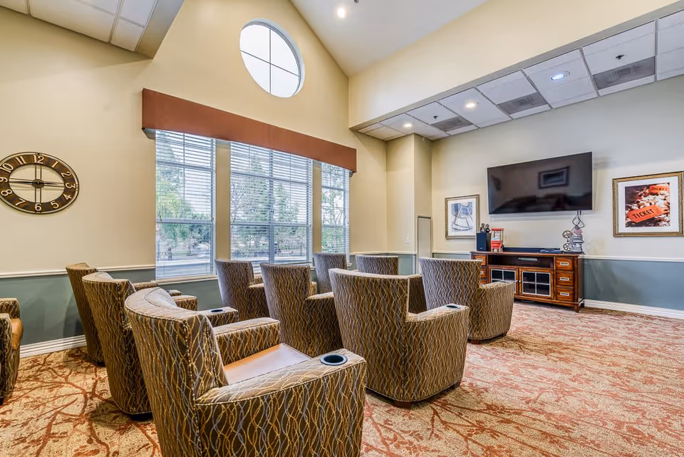 A cozy media room with multiple patterned armchairs arranged in rows facing a large flat-screen TV mounted on the wall. The room features a high ceiling with a round window above three large rectangular windows with blinds, a wall clock, framed pictures, and a wooden cabinet beneath the TV holding a popcorn machine and decorative items.