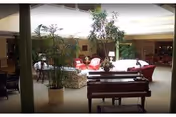 A spacious living room area with several seating arrangements including red armchairs and floral-patterned sofas. There are large potted plants placed around the room, and a piano is positioned in the foreground. The room is well-lit with natural light coming from a skylight above.
