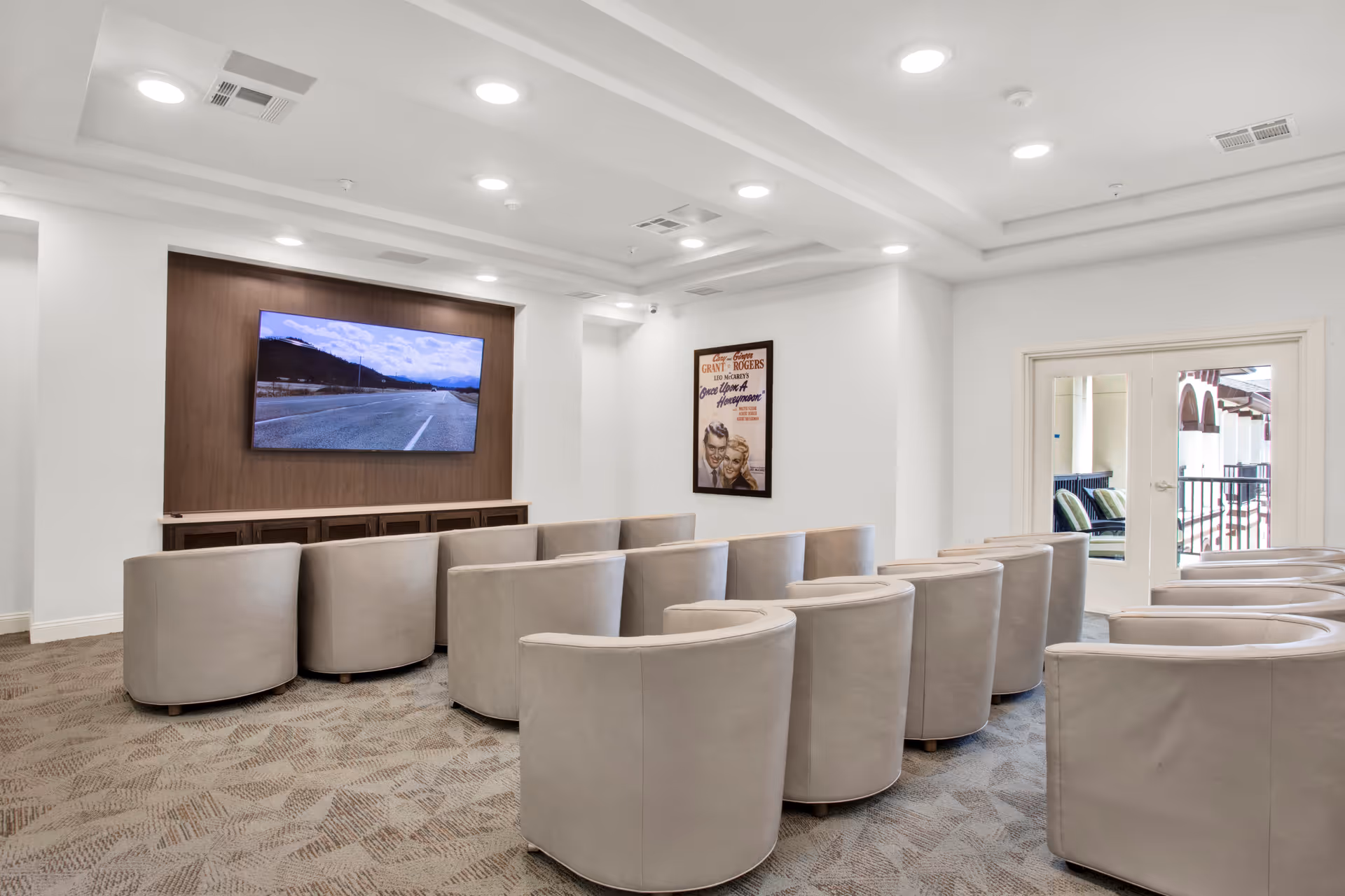 A modern media room with two rows of curved beige chairs facing a large flat-screen TV mounted on a wooden panel. The room has white walls, recessed ceiling lights, and a framed vintage movie poster on one wall. Double glass doors lead to an outdoor balcony with seating visible.