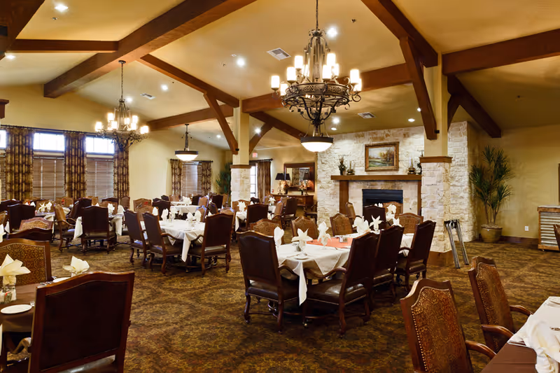 A spacious dining room with multiple tables covered in white tablecloths and neatly folded napkins. The room features wooden chairs, large windows with curtains, chandeliers hanging from a high ceiling with exposed wooden beams, and a stone fireplace with a painting above it. There are plants and decorative items around the room, creating a warm and inviting atmosphere.