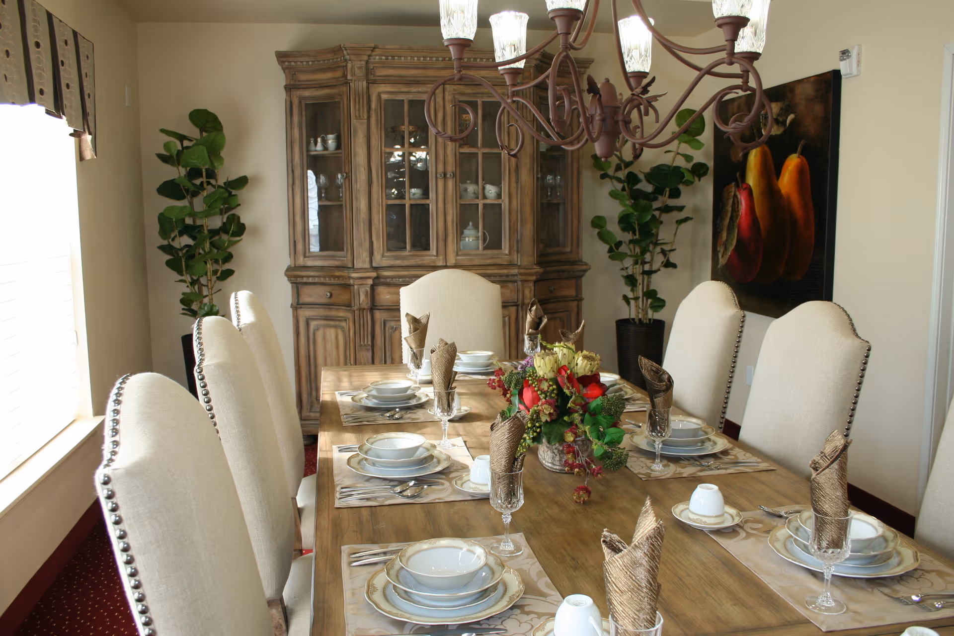 A well-lit dining room with a wooden dining table set for eight people. The table is adorned with plates, bowls, glasses, and napkins folded in an elegant style. A floral centerpiece decorates the middle of the table. Behind the table is a wooden china cabinet with glass doors displaying dishes. Two potted plants are placed on either side of the cabinet, and a painting of pears hangs on the wall to the right. A chandelier with multiple lights hangs above the table.