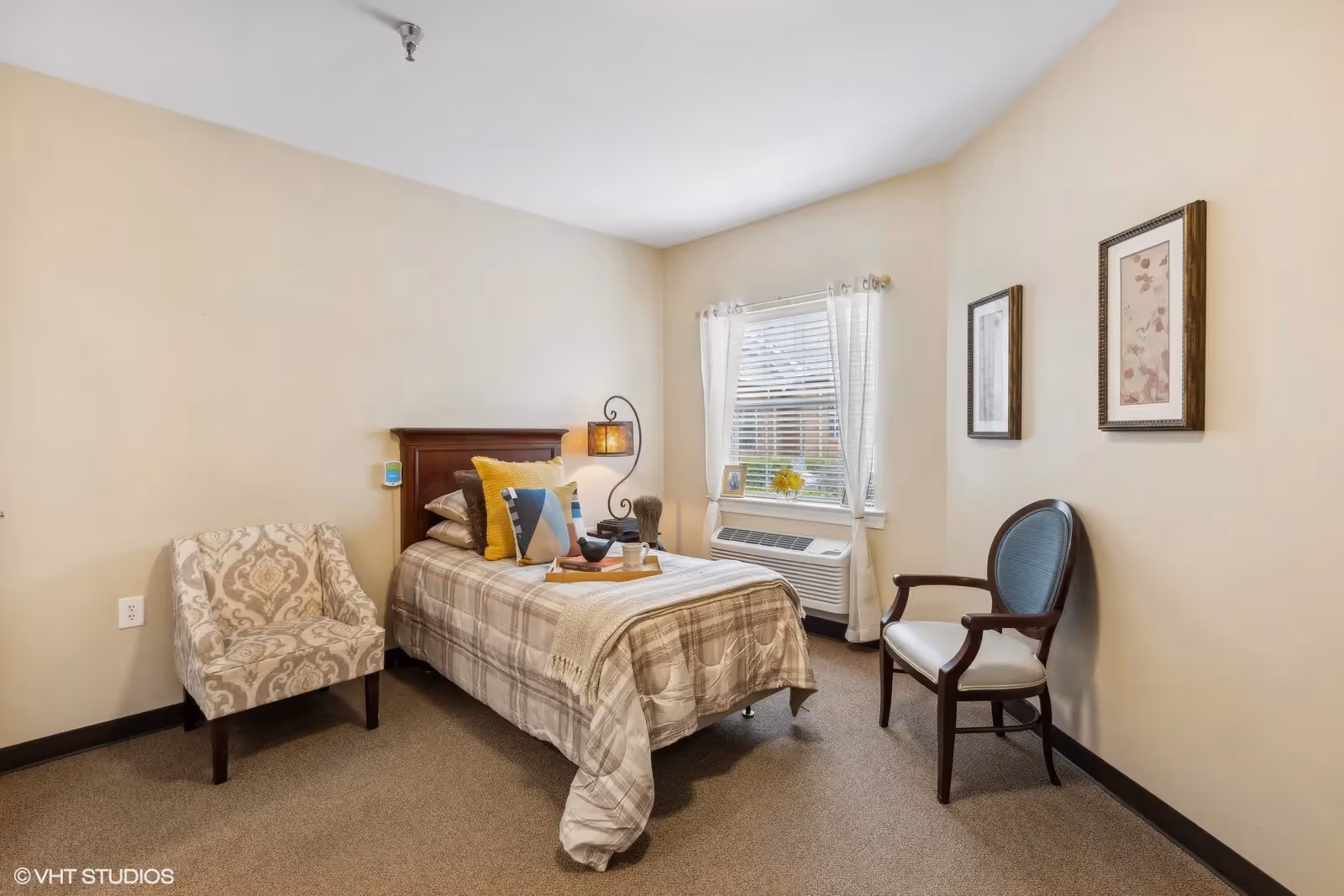 A cozy bedroom with a single bed covered in a plaid blanket and multiple pillows. There is a wooden headboard with a decorative lamp attached. Next to the bed is a patterned armchair, and on the opposite side, a wooden chair with a cushioned seat. A window with white curtains lets in natural light, and two framed pictures hang on the beige walls.