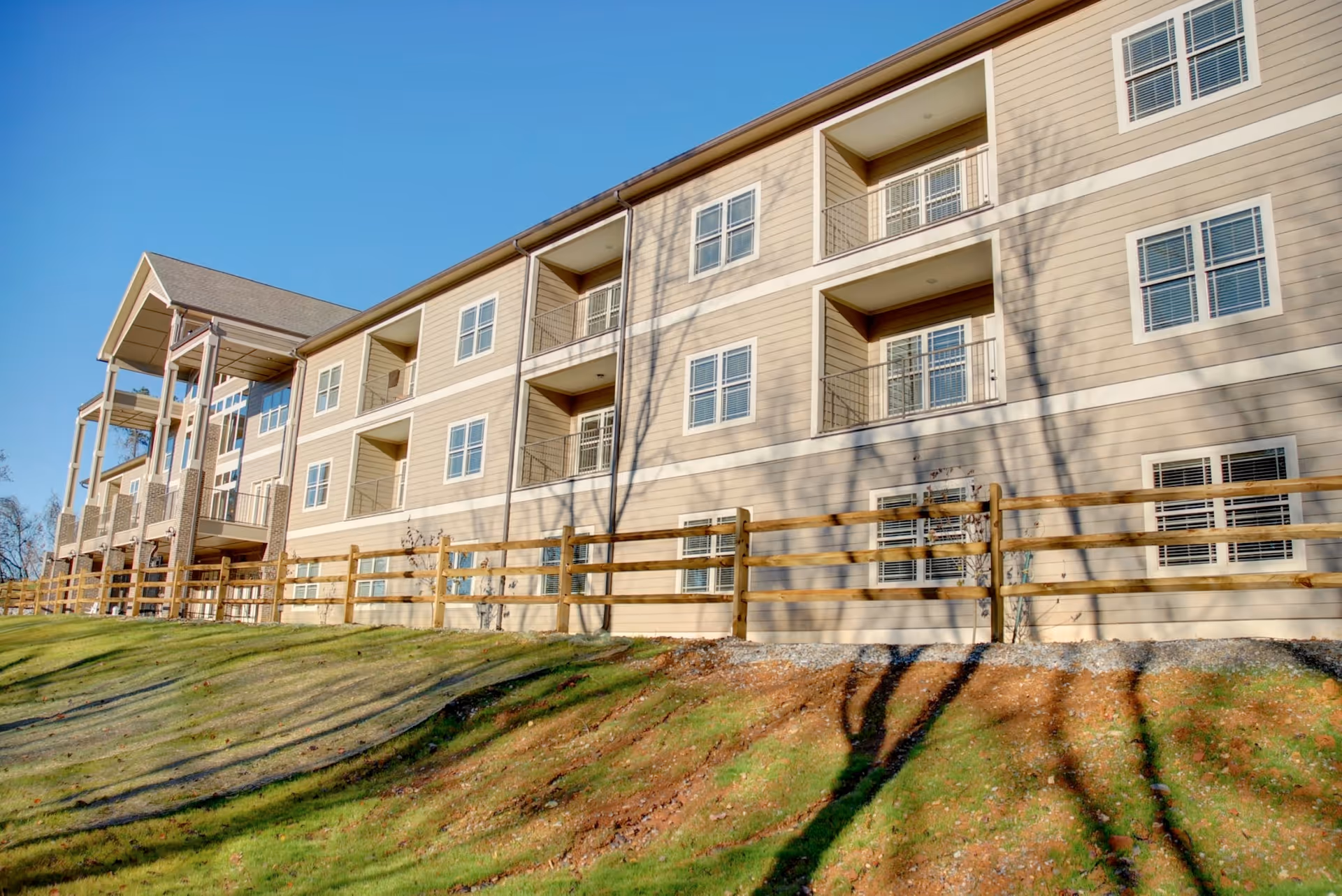 Exterior view of a multi-story senior living facility building with beige siding, multiple windows, balconies, and a wooden fence in front. The sky is clear and blue, and shadows of trees are cast on the building and grassy slope.