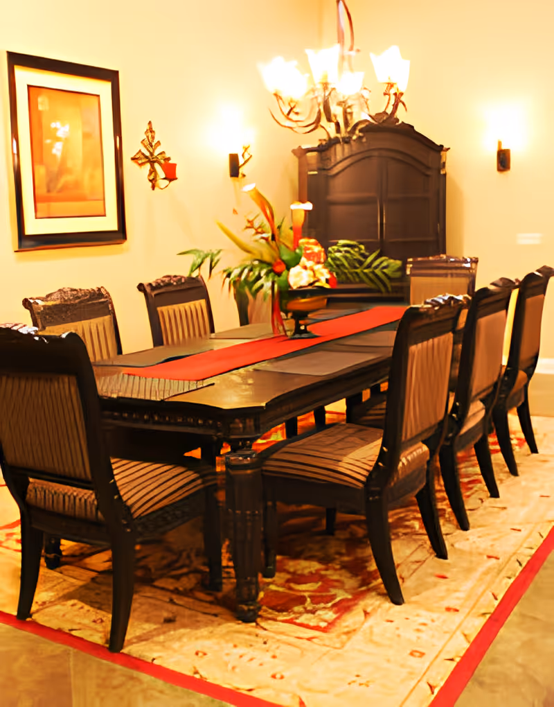 A formal dining room with a long dark wooden table surrounded by eight matching chairs with striped upholstery. The table has a red runner and a floral centerpiece. The room is warmly lit by a chandelier and wall sconces. There is a framed picture on the wall and a dark wooden cabinet in the background. The floor has a patterned area rug with red accents.