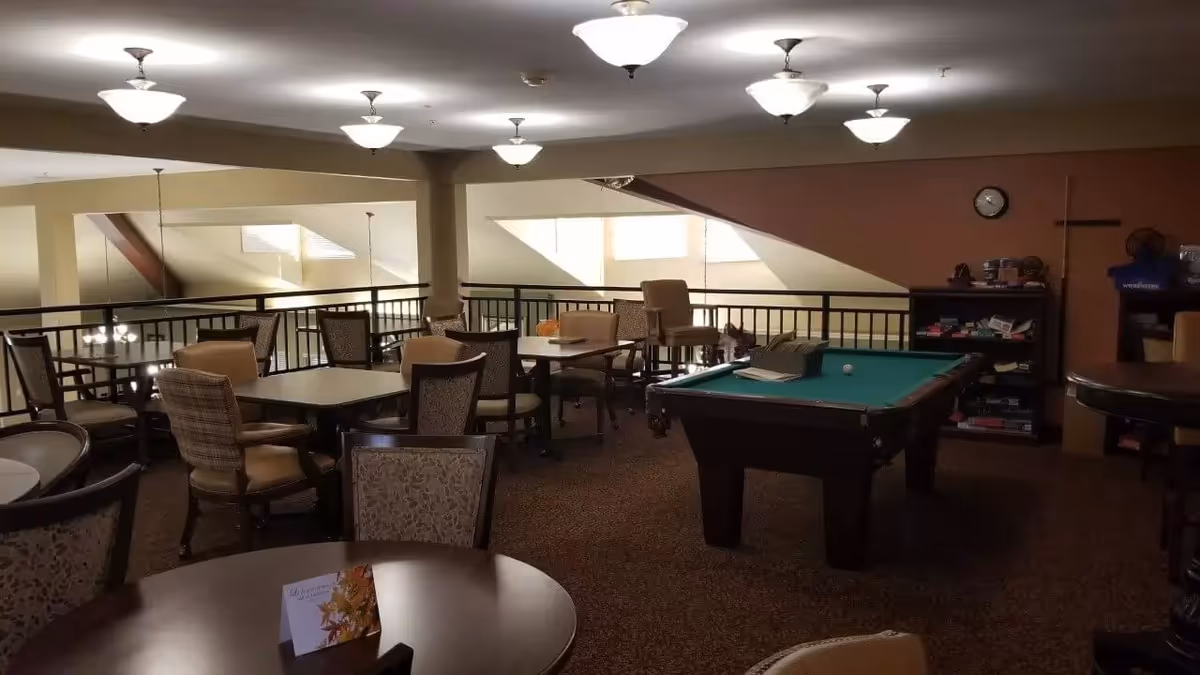 Interior view of a senior living facility common area with multiple tables and chairs arranged for seating. A pool table is positioned on the right side of the room, and a bookshelf with games and items is visible against the far wall. The room has soft lighting from ceiling fixtures and windows letting in natural light.