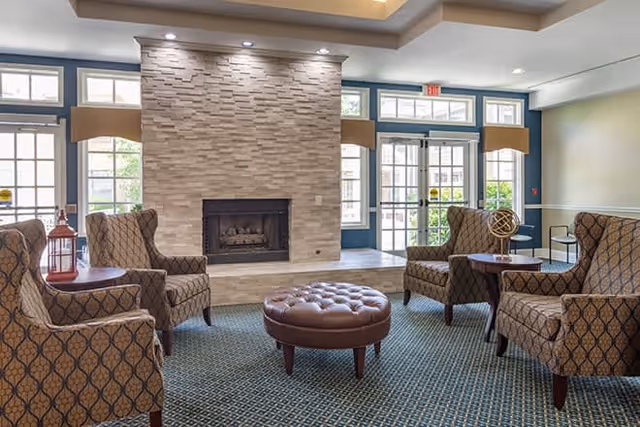 A cozy living room area with four patterned armchairs arranged around a round tufted leather ottoman. The room features a modern stone fireplace with recessed lighting above it, large windows and glass doors letting in natural light, and a carpeted floor with a geometric pattern.