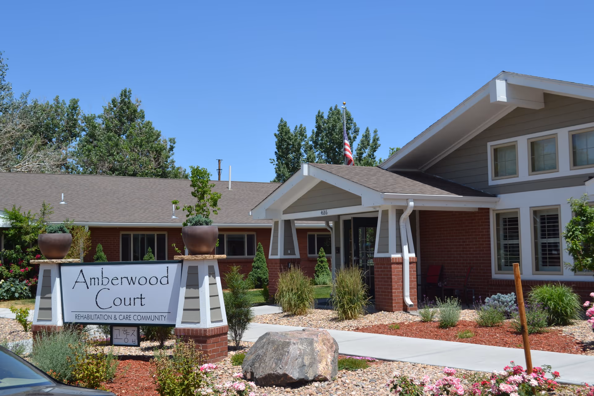 Exterior view of Amberwood Court, a rehabilitation and care community building with a brick and siding facade, a covered entrance, landscaped garden with flowers and shrubs, and an American flag on the roof.