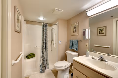 A clean and accessible bathroom featuring a walk-in shower with a floral shower curtain, a white toilet, a sink with a countertop, a large mirror above the sink, and two blue towels hanging on a towel rack. The walls are beige with framed artwork, and there are grab bars installed near the shower and toilet for safety.