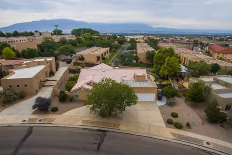 Aerial view of a residential neighborhood with single-story houses, trees, and a mountain range in the background under a cloudy sky.