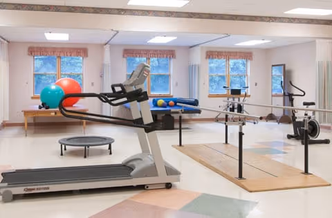 A physical therapy or rehabilitation room with exercise equipment including a treadmill, parallel bars for walking practice, a small trampoline, exercise balls, and a stationary bike. The room has large windows with curtains and a clean, spacious layout.