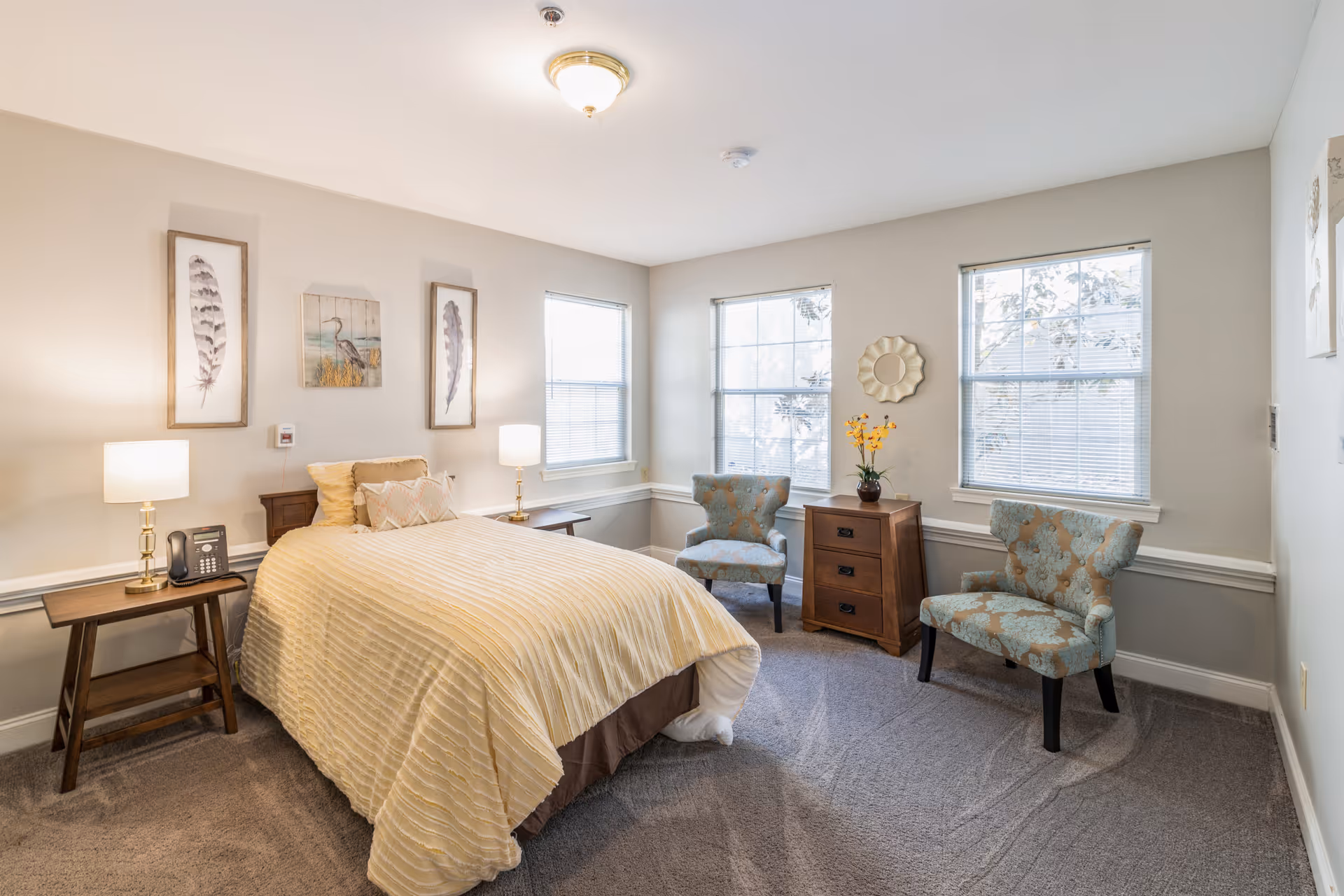 A bright and cozy bedroom with a single bed covered in a beige striped comforter, two bedside tables with lamps, two patterned armchairs, a wooden chest of drawers with a vase of yellow flowers, and three framed artworks on the walls. The room has three large windows with blinds and light gray walls.