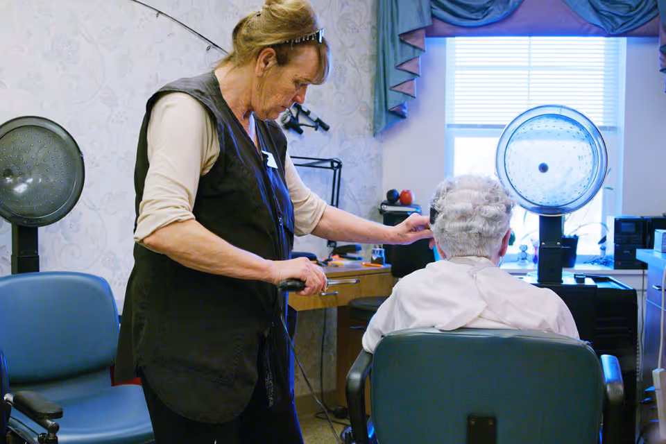 A hairdresser styling the hair of an elderly woman seated in a salon chair in a room with hair drying equipment and a window with blue curtains.