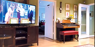 A cozy living room area featuring a large flat-screen TV on a dark wooden stand, an upright piano with a red cushioned bench, framed pictures on the wall, and an open door leading to another room.