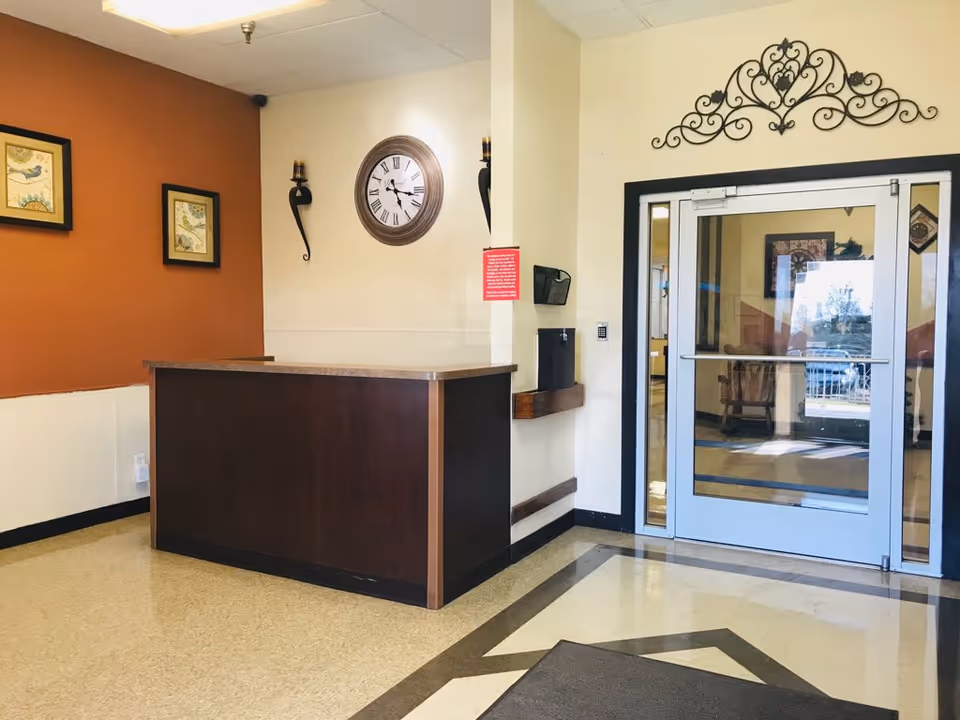 Reception area with a dark wooden desk, a wall clock showing 10:10, two framed pictures of birds on an orange wall, decorative wall sconces, and a glass door entrance with a metal frame. The floor has a geometric pattern near the entrance.