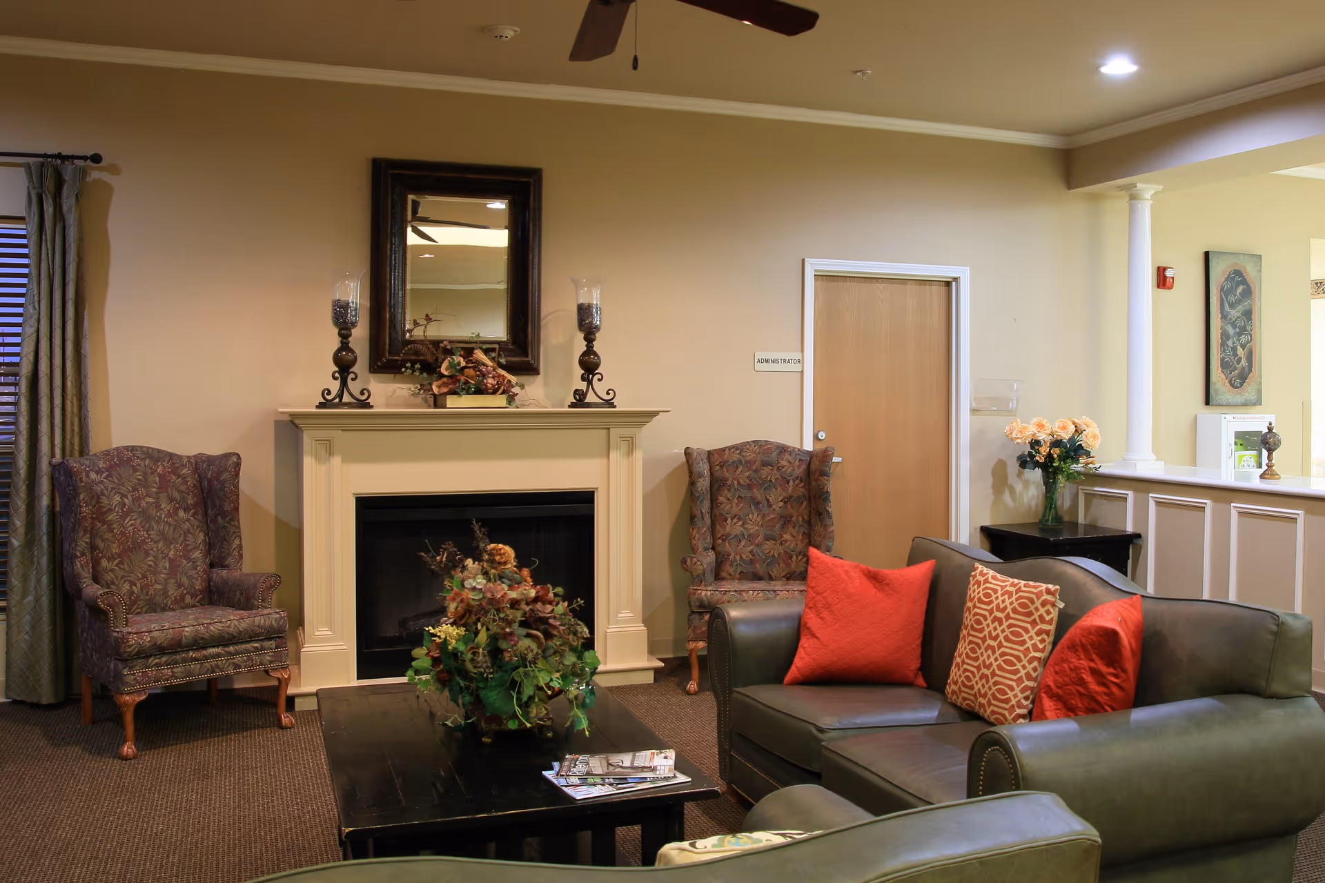 A cozy living room area with a dark green leather sofa adorned with red and patterned cushions, two patterned armchairs, a black coffee table with a floral centerpiece and magazines, a white fireplace with decorative candle holders and a mirror above it, beige walls, and a door labeled 'Administrator'.