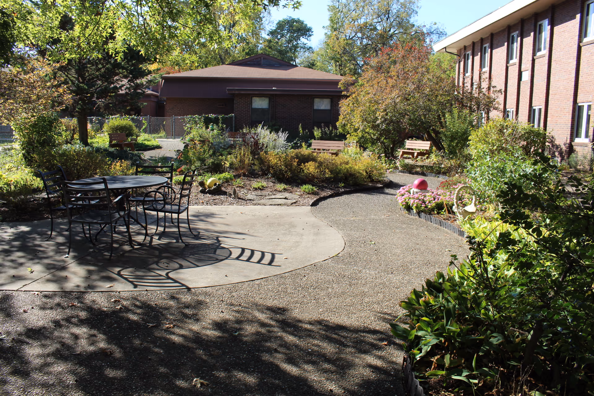 Outdoor garden area at The Grove Health and Rehab featuring a paved walkway, metal table with chairs, benches, various plants, and trees with a brick building in the background.