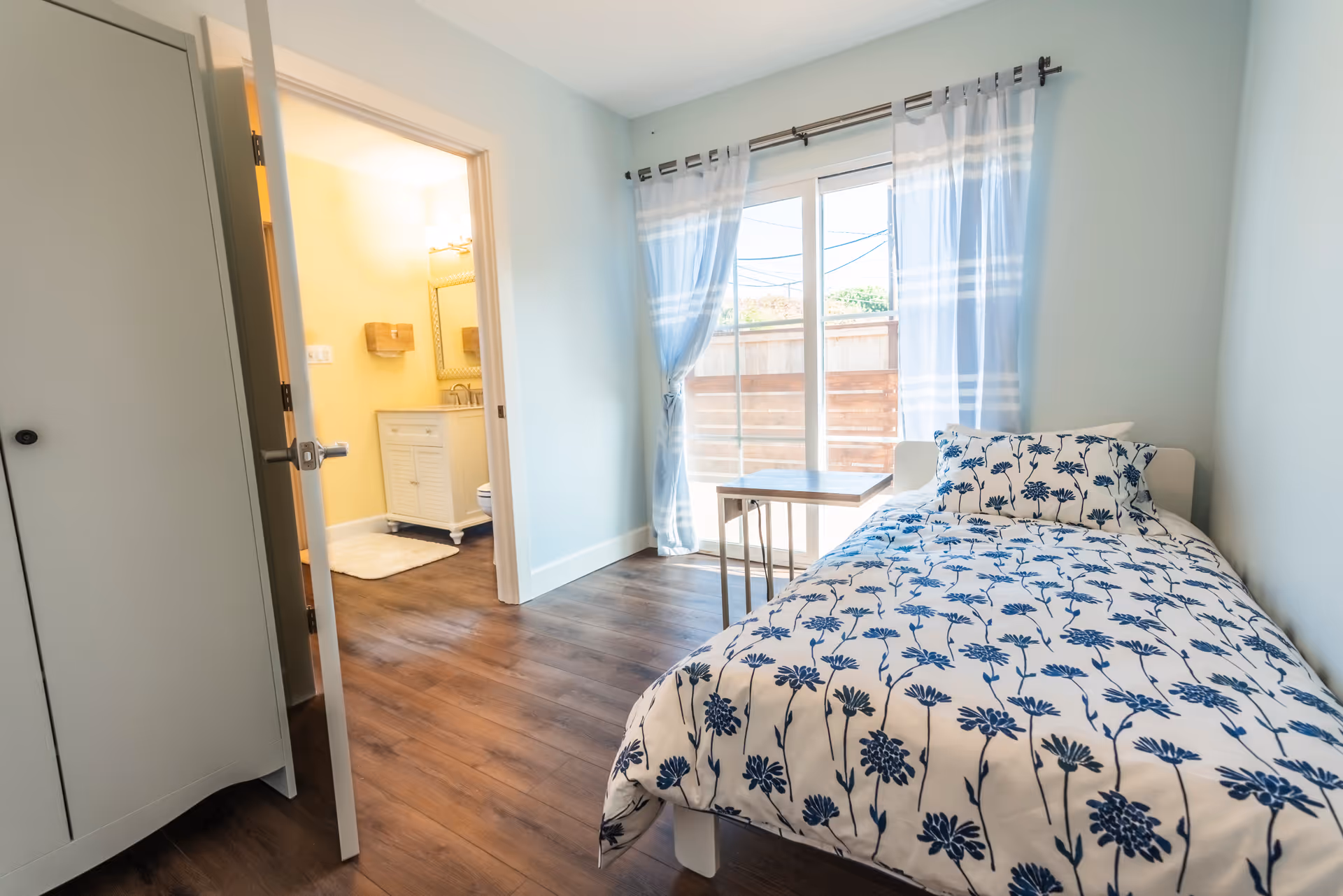 A bright bedroom with a single bed covered in white bedding with blue floral patterns. Next to the bed is a small table. The room has wooden flooring and light blue walls. A large sliding glass door with sheer white curtains lets in natural light. An open door reveals a bathroom with a white vanity and a mirror.