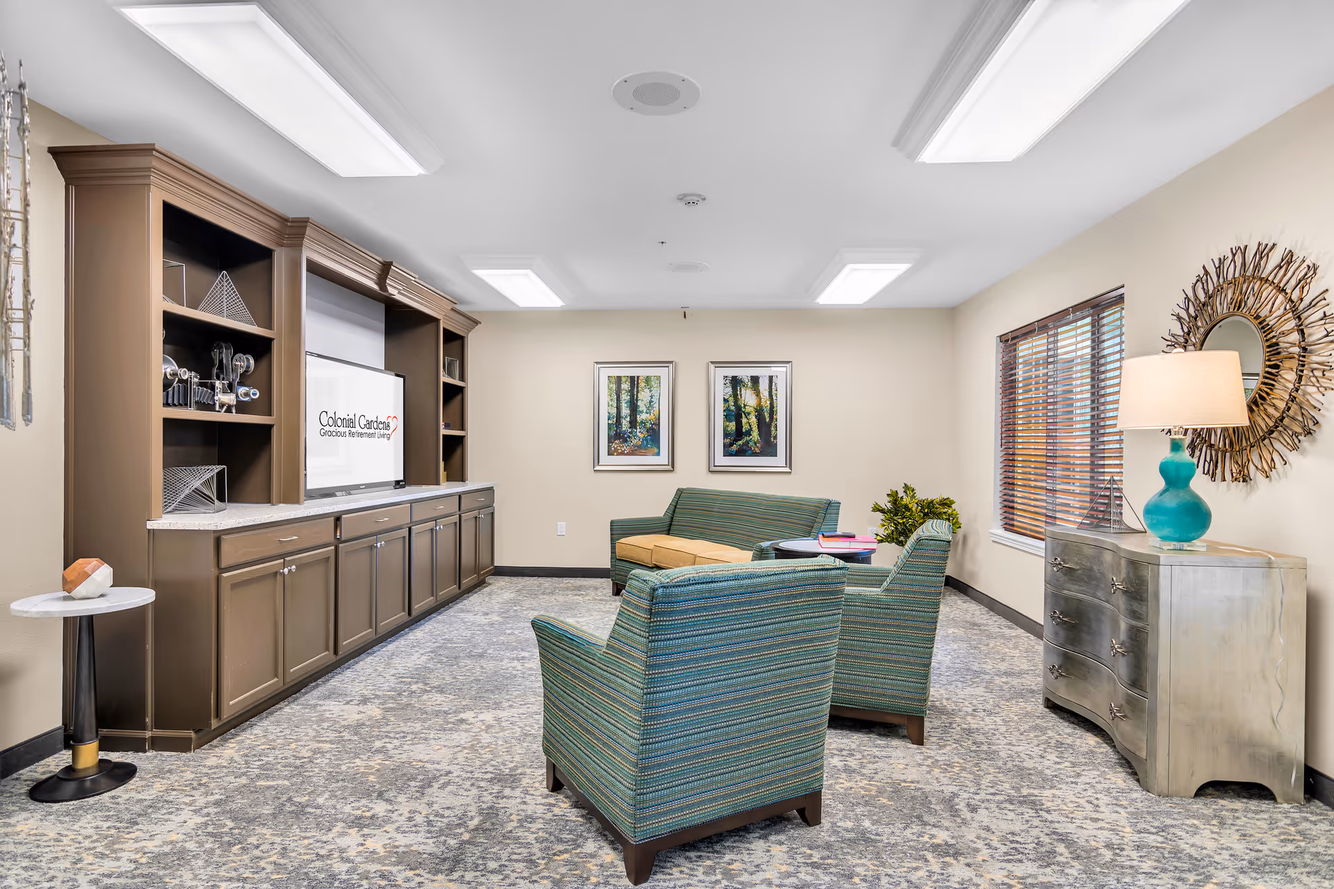 Communal living room with striped armchairs, a built-in TV cabinet displaying 'Colonial Gardens Gracious Retirement Living', artwork, and a sideboard with a lamp.