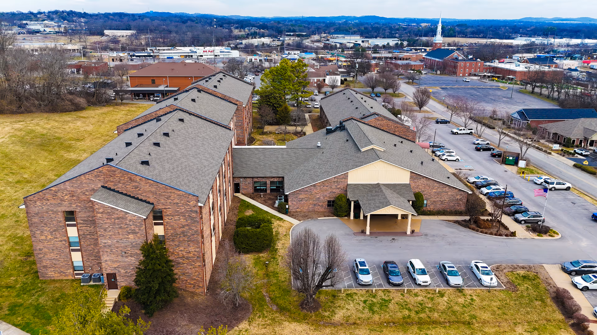 Aerial view of a brick senior living complex with an entrance canopy, parking lot, and surrounding town.