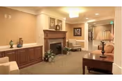 A cozy interior common area with beige walls and carpeted floor. The room features a wooden fireplace mantel with decorative items on top, a framed picture on the wall, a table lamp on a wooden side table, and comfortable seating including a sofa and armchairs. The space is softly lit with ceiling lights and has a calm, welcoming atmosphere.