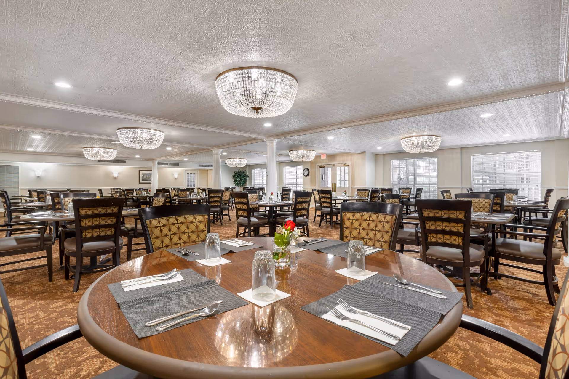 A spacious dining room with multiple round and square wooden tables set with placemats, utensils, and glasses. The chairs have patterned upholstery and the room is well-lit with large crystal chandeliers and recessed ceiling lights. Windows along the walls allow natural light to enter, and there are white columns and light-colored walls throughout the room.