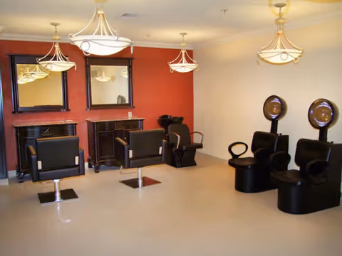 Interior view of a salon area with three black salon chairs in front of mirrors on a red wall and three black hair drying chairs with hooded dryers on the right side. The room has beige flooring and three ceiling light fixtures.
